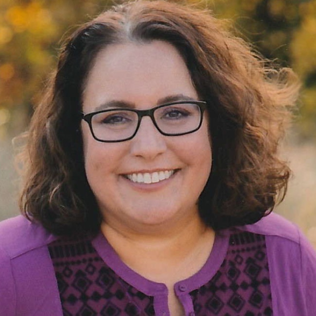 A person smiling and wearing glasses with a necklace.