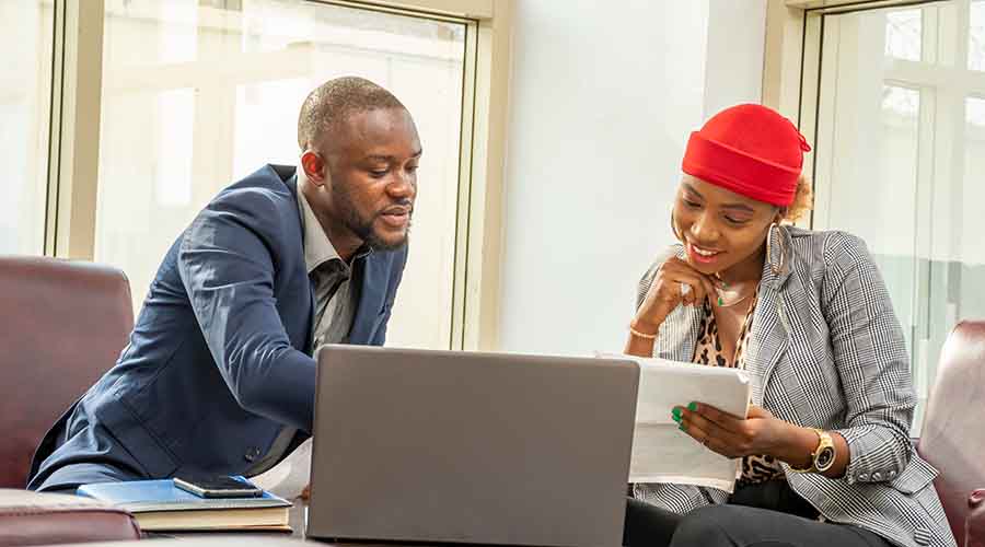 A man with a shaved head and beard wearing a blue blazer reviews paperwork with a woman wearing a red hat and checkered blazer. They sit in front of a desk that has a laptop, books, and a mobile phone on top of it.